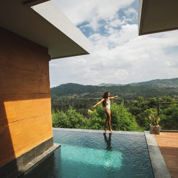 Slim beautiful woman in white swimsuit walking by edge of infinity swimming pool with amazing jungle