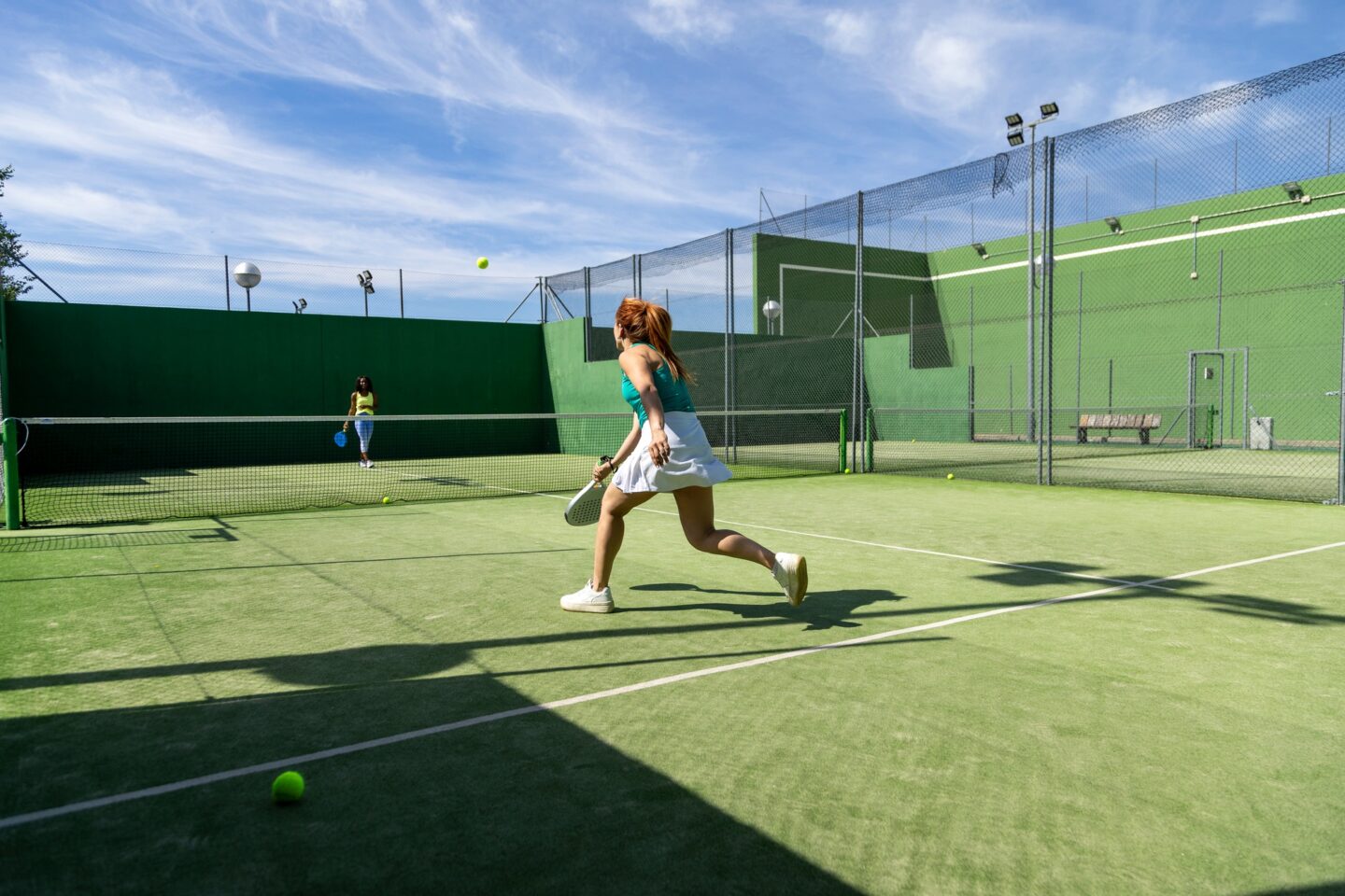 Two women playing padel on an outdoor court, one chasing the ball in mid-action.